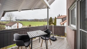 A covered outdoor patio with a table and chairs, a view of a green field, and a building in the background. - Holiday Apartment in the Countryside with Mountain View, Balcony and Wi-Fi (Wolpertswende)