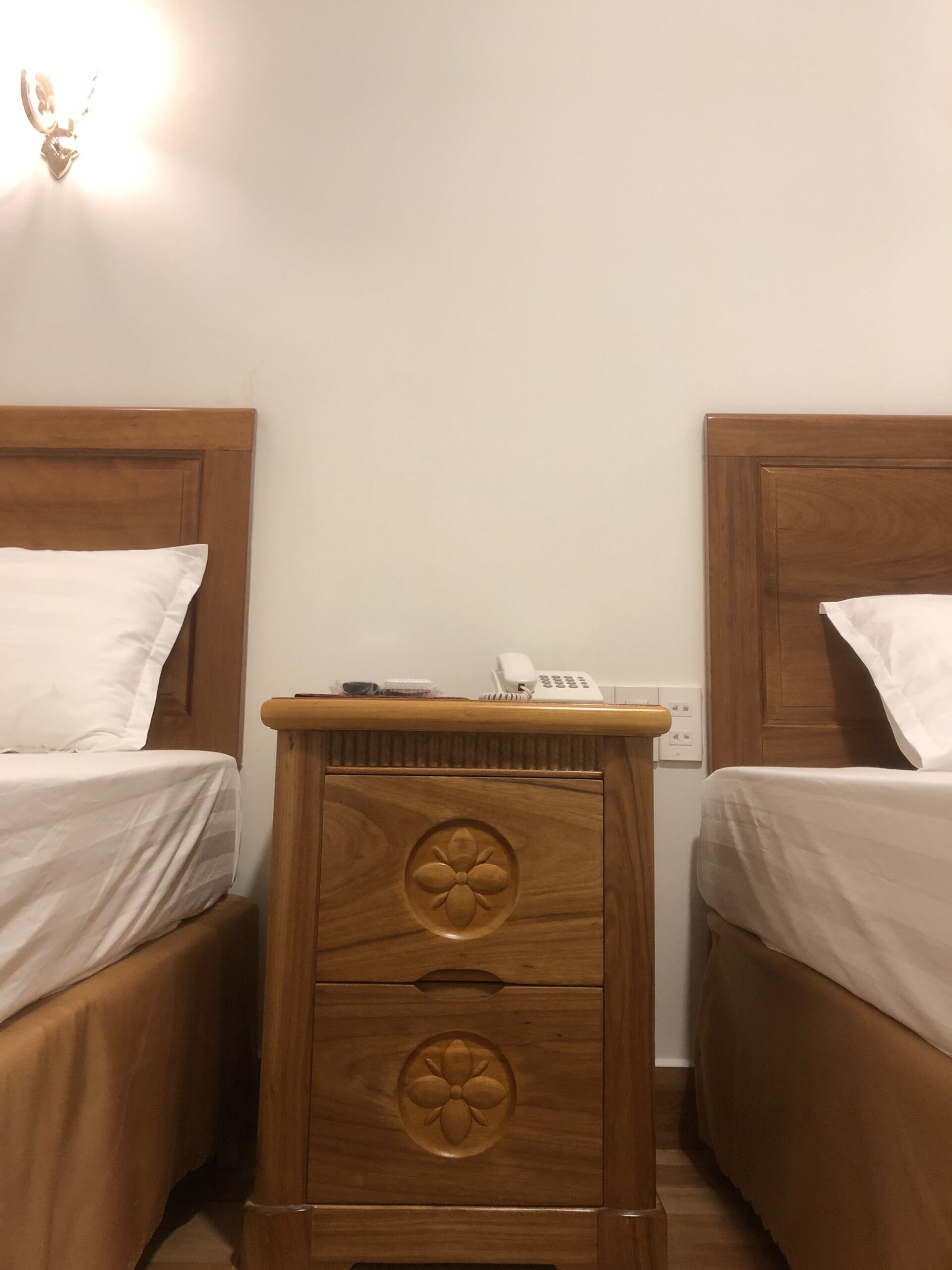 A room with two wooden beds, each with white bedding. There is a wooden nightstand between the beds with a phone and a small item on top.