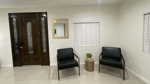 A room with a dark wooden door, two black leather chairs, a small round table with a plant, a mirror on the wall, and a window with blinds.