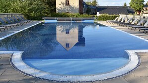 A large, clear swimming pool with a tiled edge, surrounded by lounge chairs and a building in the background. - Bridge House (Cirencester)
