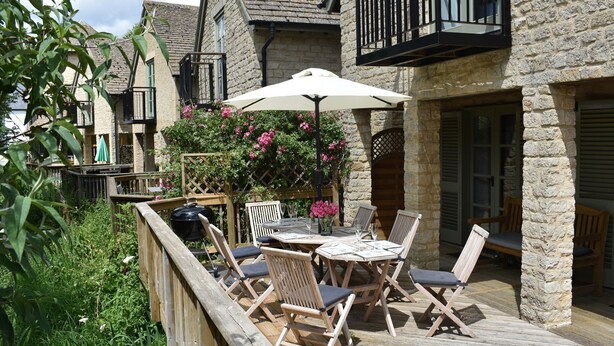 A patio area with a table and chairs, surrounded by stone walls and a wooden deck. - Bridge House (Cirencester)