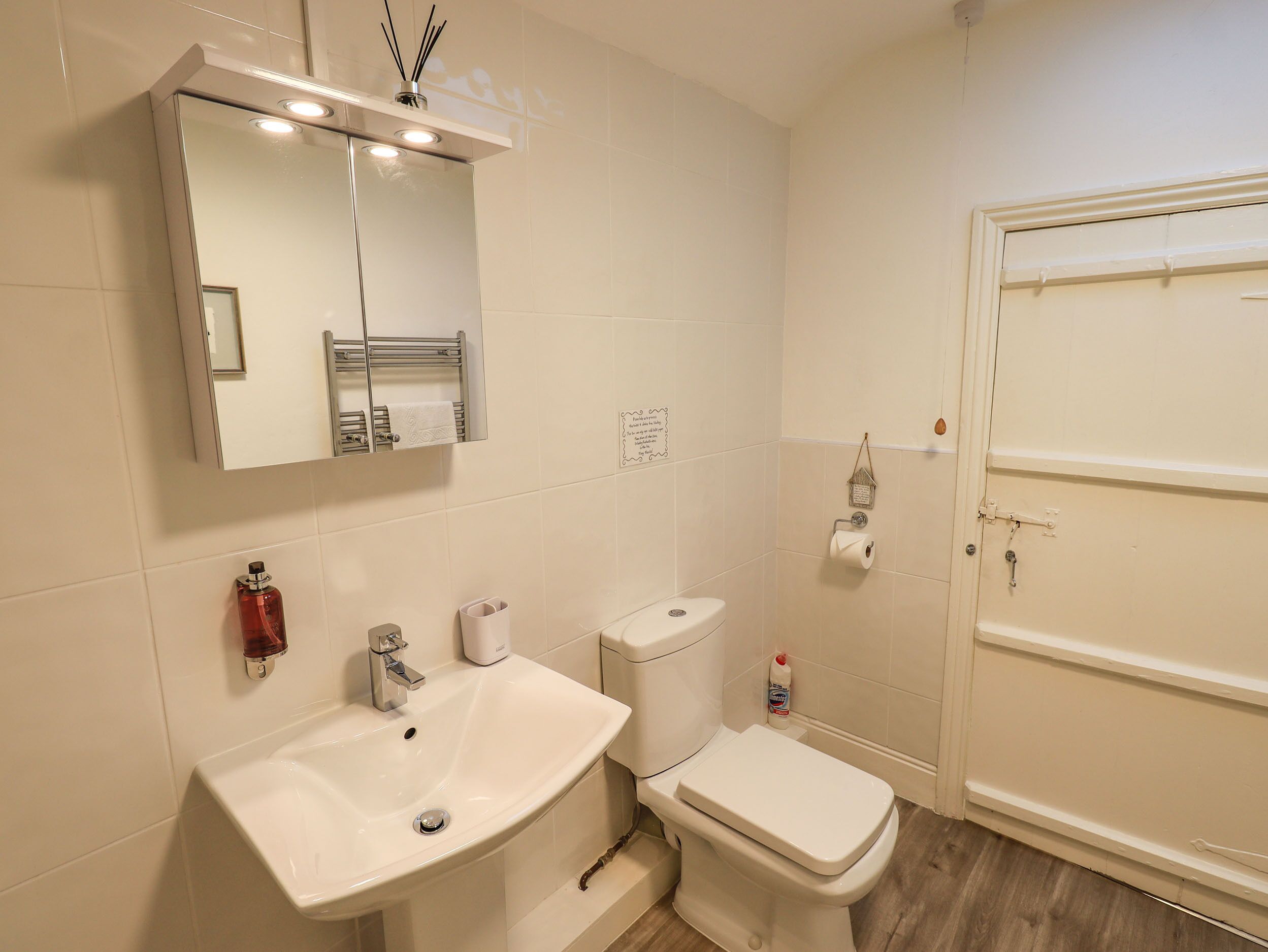 A bathroom with a white sink, toilet, and mirror. There is a towel rack above the mirror, a bottle of hand soap, and a toilet paper holder.