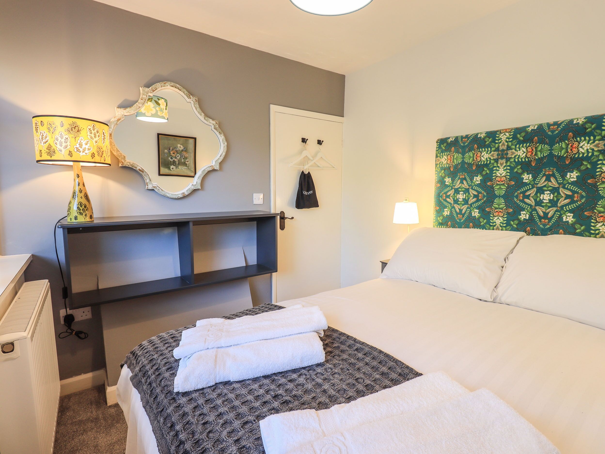 A hotel room with a bed, a desk with a lamp, a wardrobe, and a patterned wall tapestry.