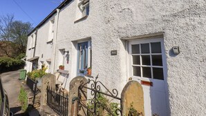 A white building with a blue door and window shutters, a small garden with potted plants, and a metal gate. - Violet Cottage (Ambleside)