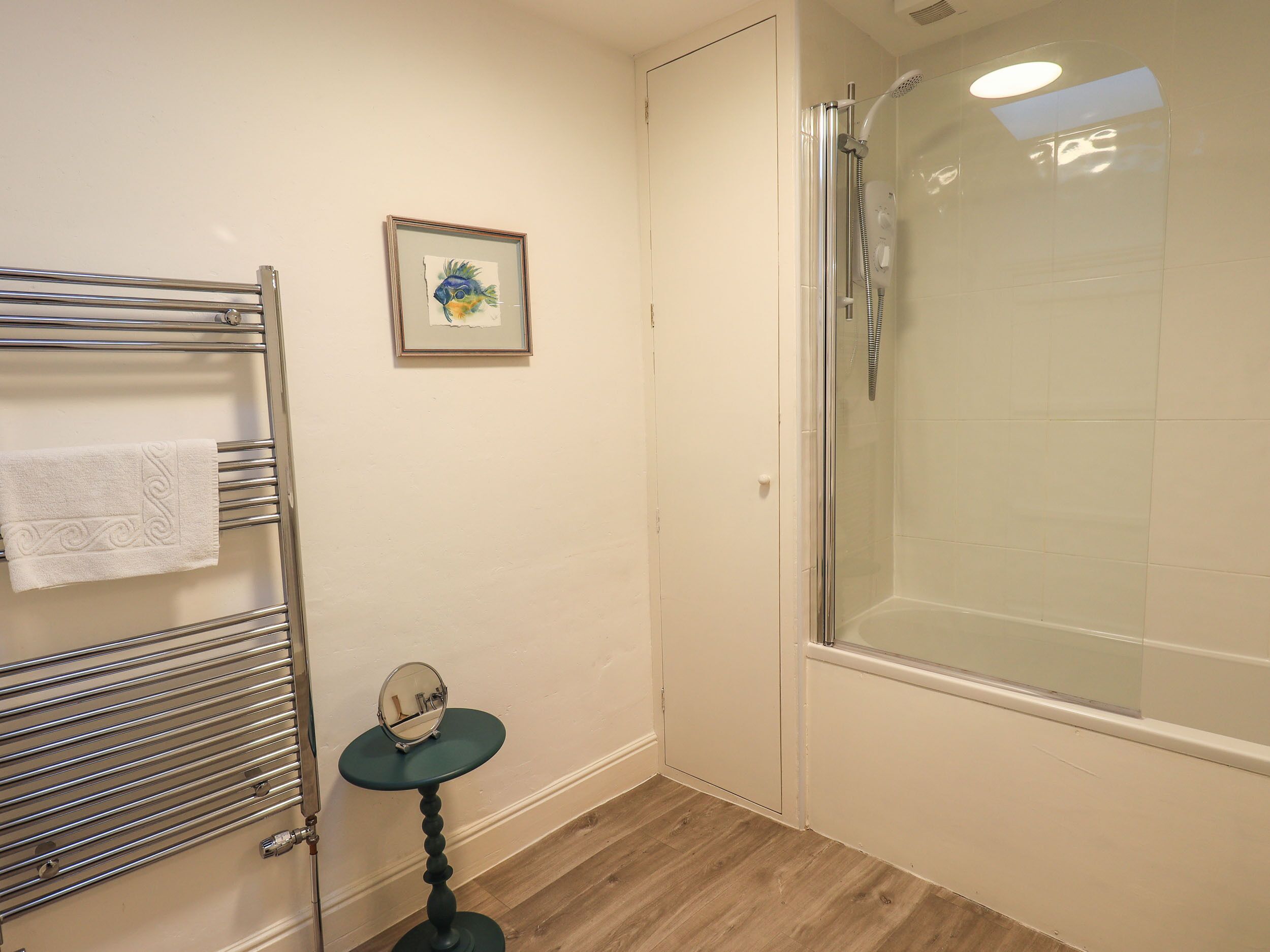 A bathroom with a glass-enclosed shower, a towel rack with a white towel, a small round table with a mirror, and a framed picture on the wall.