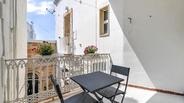 Een balkon met een witte reling, twee zwarte stoelen en bloempotten.