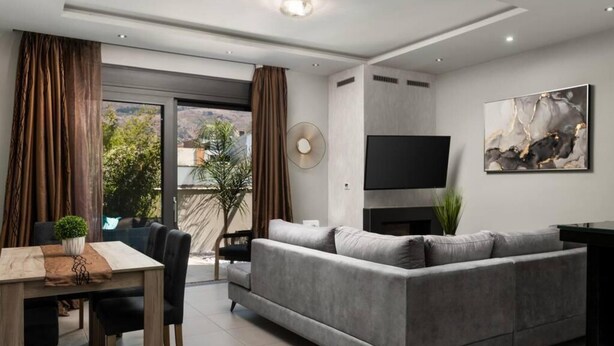 Ein modernes Wohnzimmer mit grauem Sofa, einem Esstisch mit Stühlen, einem flachbildfernseher an der Wand und einem großen Fenster mit Vorhängen.