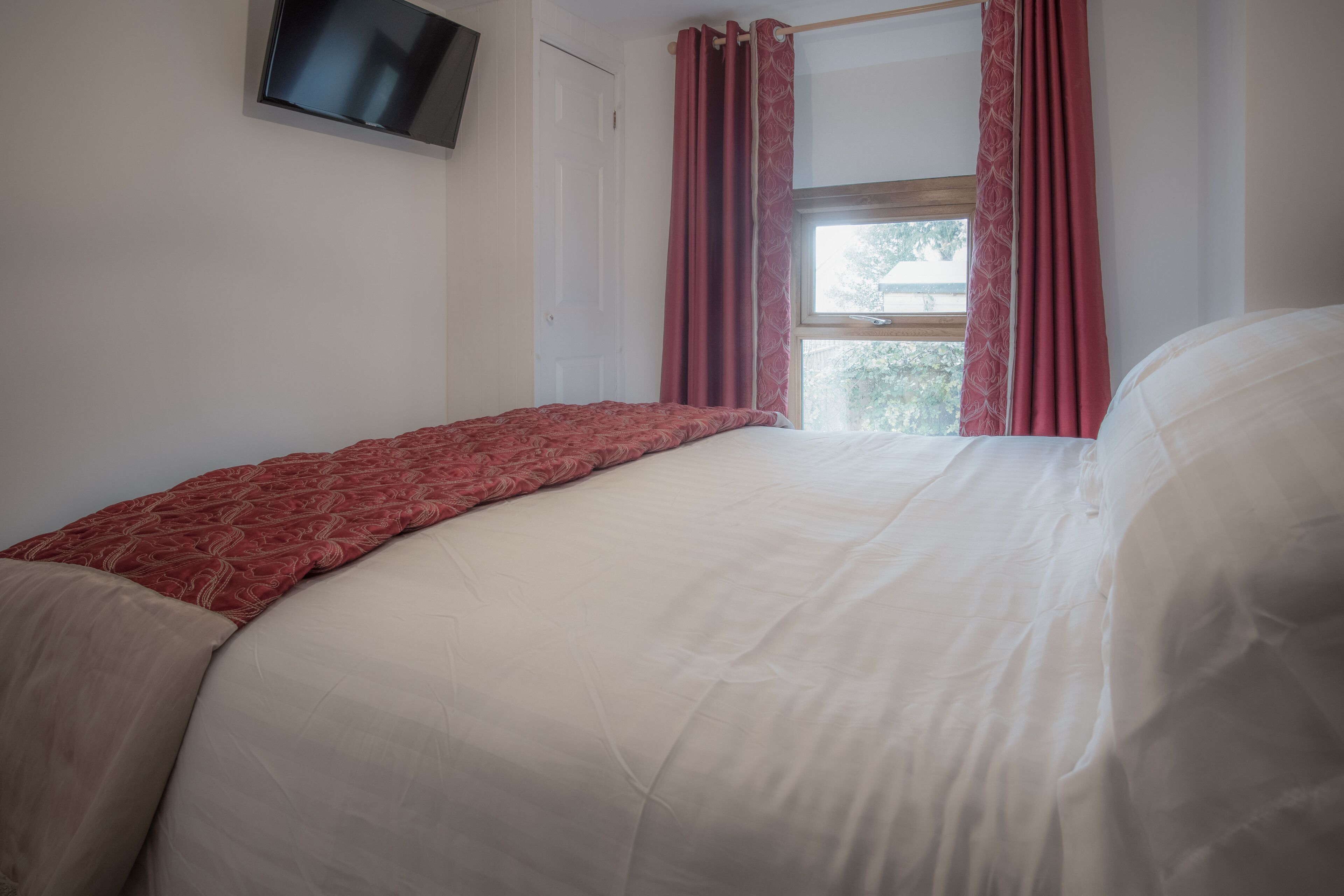 Ein Schlafzimmer mit einem Bett, einem an der Wand montierten Fernseher und einem Fenster mit Vorhängen.