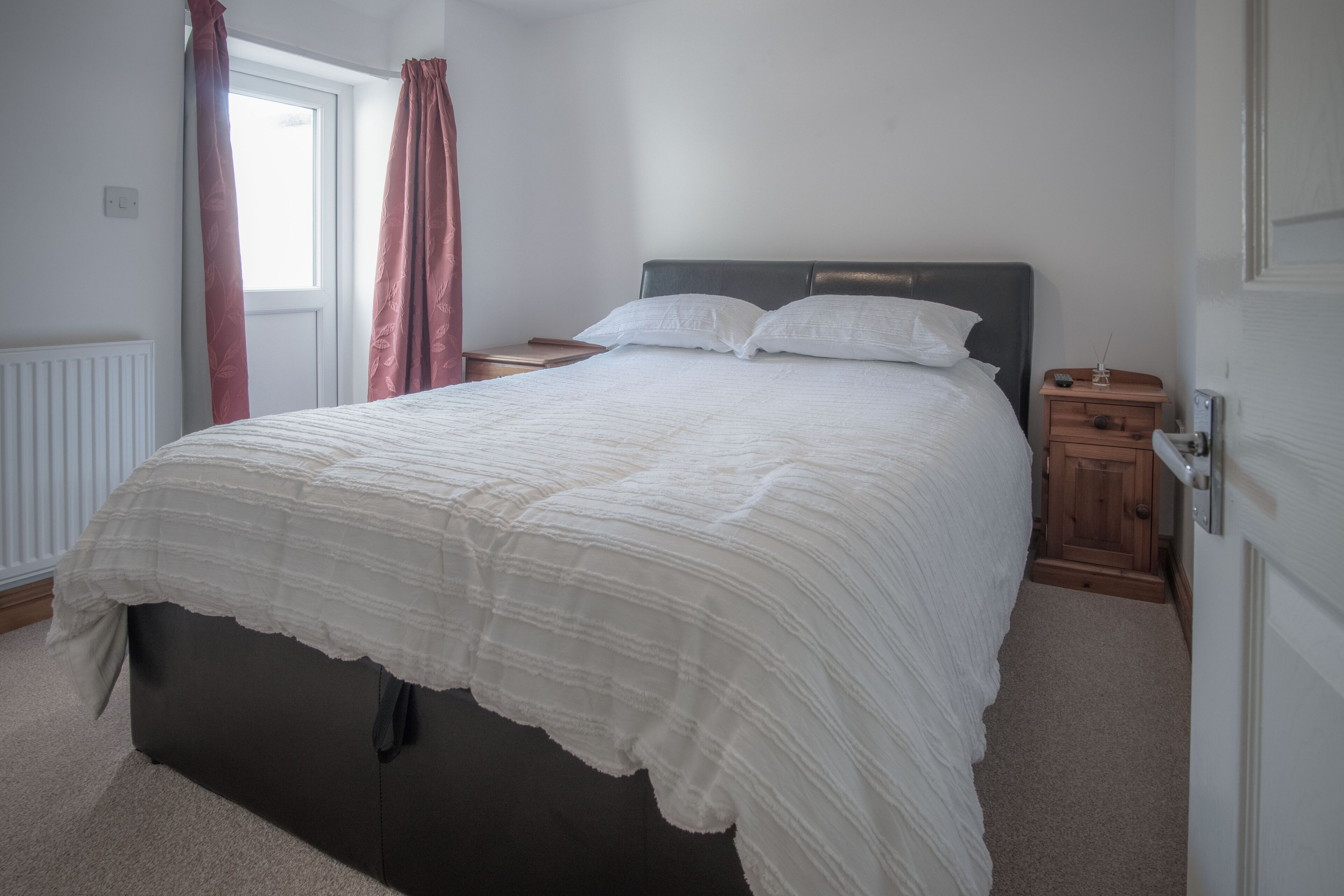Een slaapkamer met een groot bed, een nachtkastje, een houten garderobe en een raam met gordijnen.