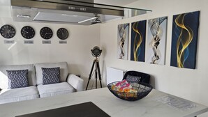 A modern kitchen with a marble countertop, a black induction cooktop, a glass coffee table, and a black chair. There are four wall clocks showing different time zones and abstract paintings above the coffee table. - Luxury Small Cozy House With Lake View (Doncaster)