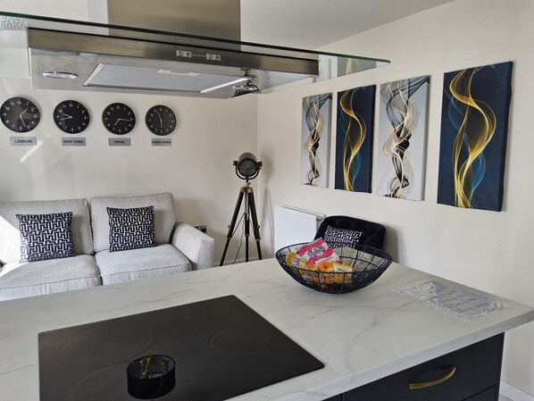 A modern kitchen with a marble countertop, a black induction cooktop, a glass coffee table, and a black chair. There are four wall clocks showing different time zones and abstract paintings above the coffee table. - Luxury Small Cozy House With Lake View (Doncaster)