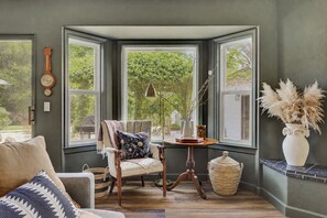 A room with a bay window, a wooden chair, a table with a lamp, and a vase with dried plants. - Château Vigne: Luxe retreat, Hot Tub, plunge pool, Outdoor movies, game room, EV (Atascadero)
