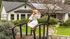 A woman in a white dress and hat standing on a wooden bridge in a garden with a house in the background. - Château Vigne: Luxe retreat, Hot Tub, plunge pool, Outdoor movies, game room, EV (Atascadero)