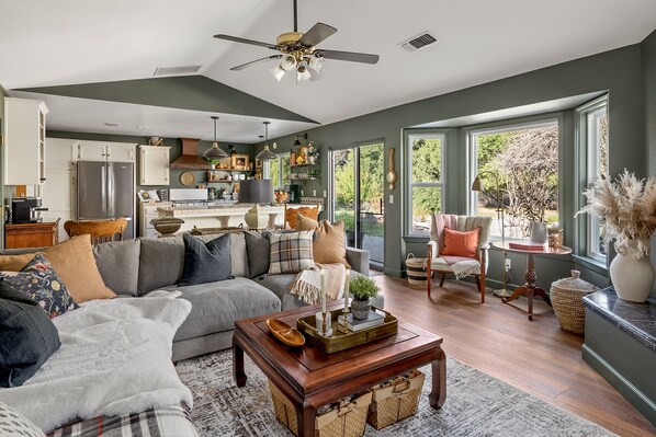 A spacious living room with a sofa, armchairs, and a coffee table. There is a kitchen in the background with a refrigerator and cabinets. - Château Vigne: Luxe retreat, Hot Tub, plunge pool, Outdoor movies, game room, EV (Atascadero)