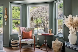 Relax in this serene corner room with a bay window overlooking a lush garden, featuring a classic armchair and a small table with a lamp.