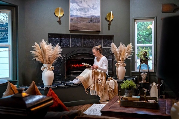 A cozy living room with a fireplace, two vases with pampas grass, and a person reading a book.