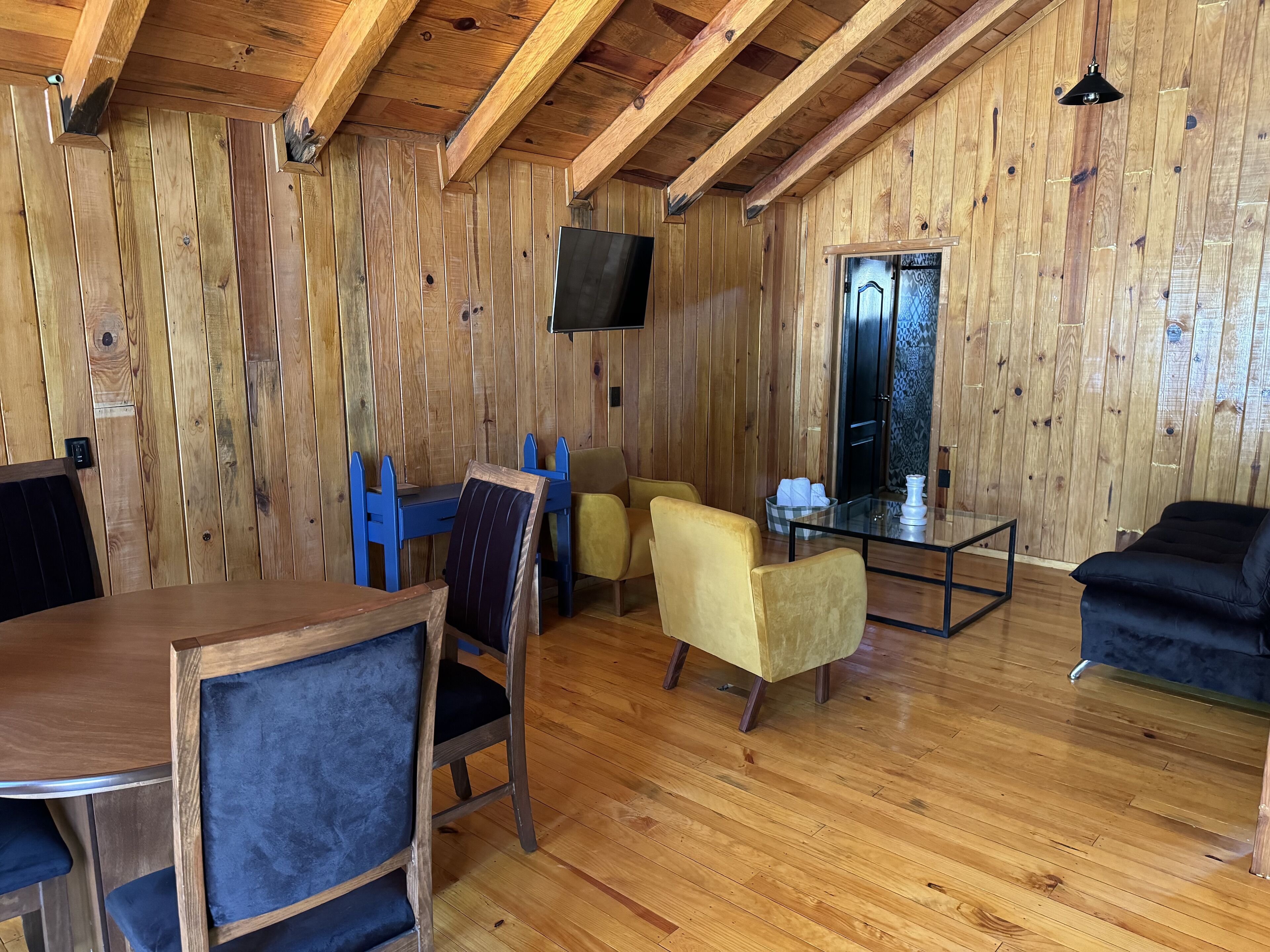 Een kamer met houten lambrisering, een eettafel, stoelen, een bank en een aan de muur bevestigde televisie.