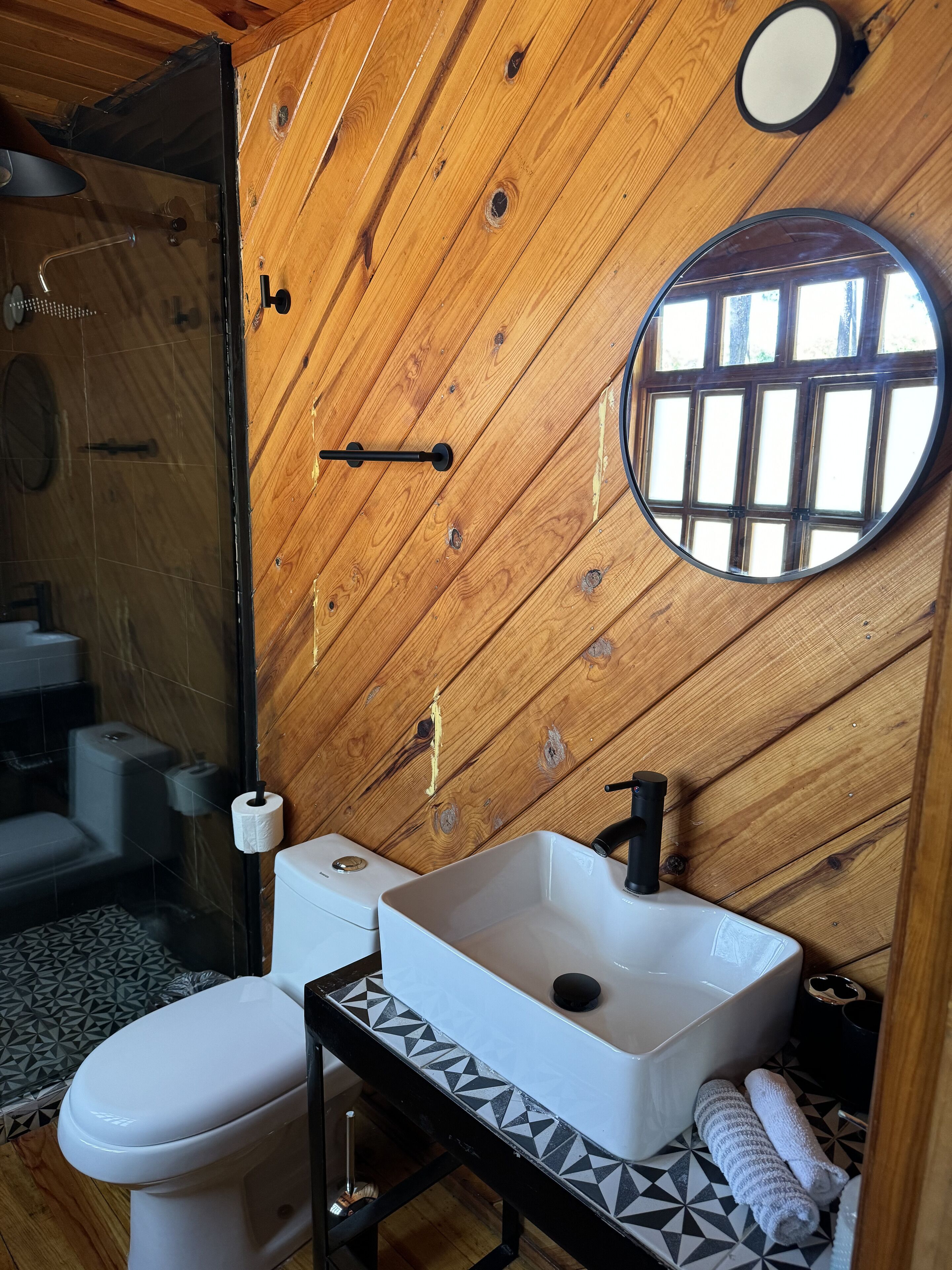 Et badeværelse med træpaneler, et hvidt toilet, en vask med en sort hane og et spejl.