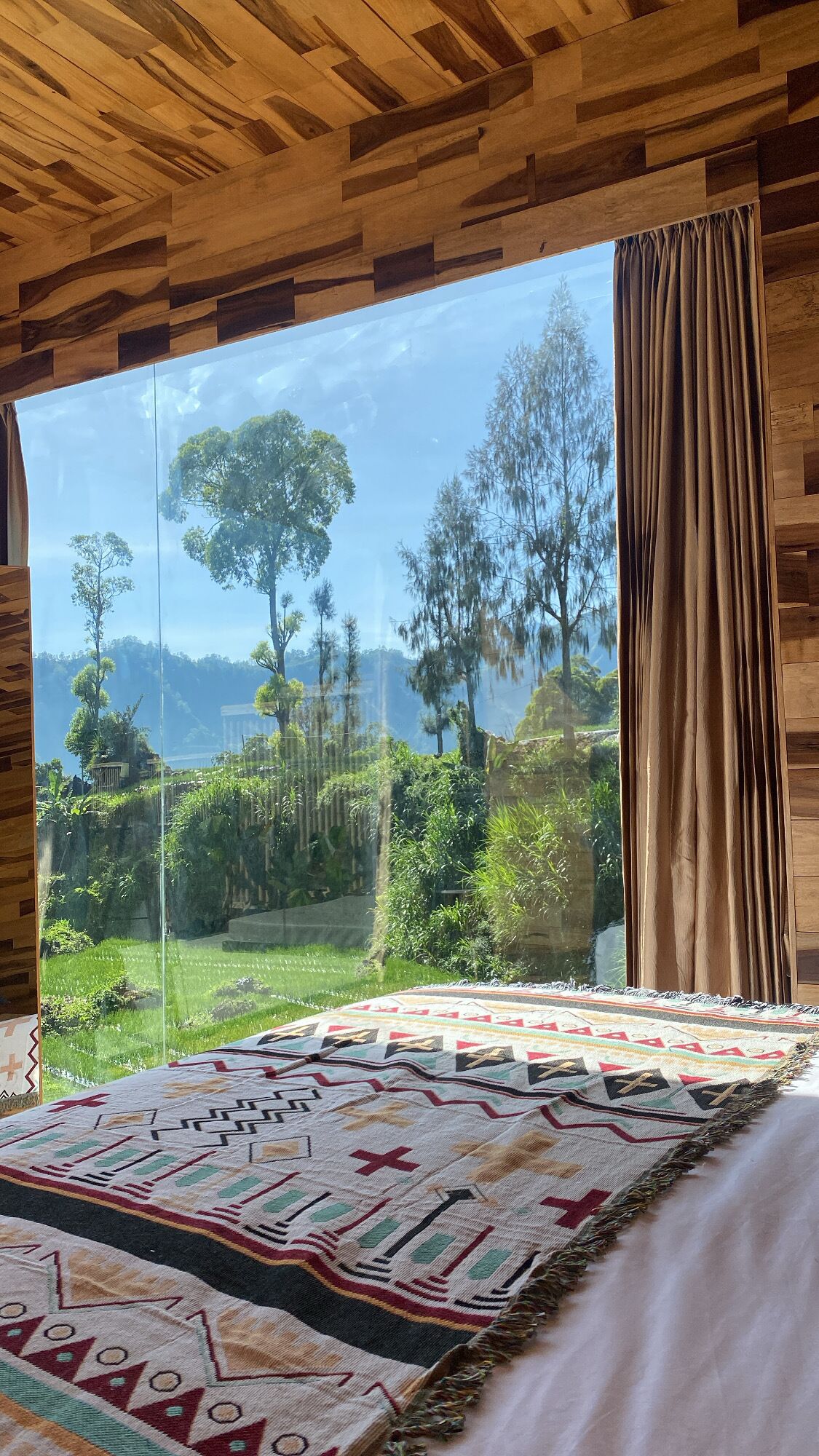 A bedroom with a large window, a bed with a patterned blanket, and wooden ceiling panels.
