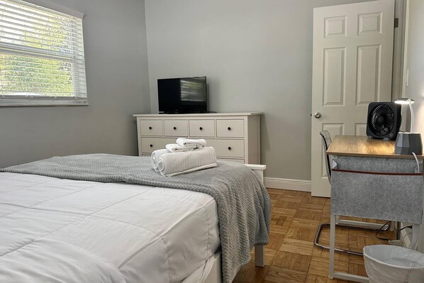 A bedroom with a bed, a dresser, a television, a fan, and a small table with a lamp.