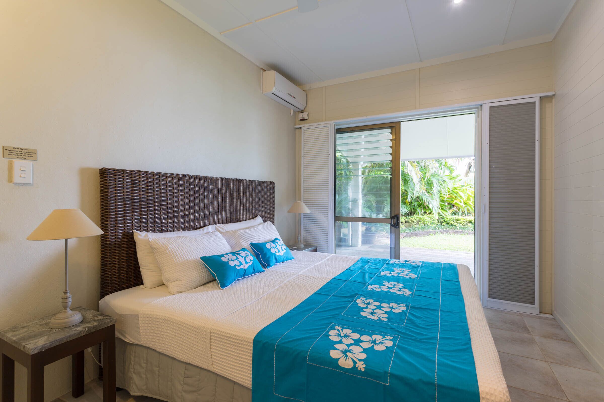 A bedroom with a bed, a nightstand, a lamp, a window with blinds, and an air conditioning unit.
