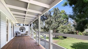 Un porche cubierto con una terraza de madera, barandillas blancas y vistas a un césped y árboles.