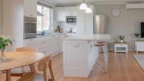 Una cocina moderna con armarios blancos, electrodomésticos de acero inoxidable, una isla central con taburetes y un comedor con mobiliario de madera.