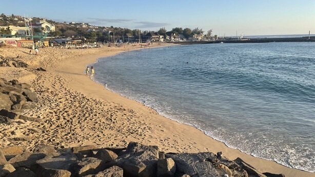 Une plage au rivage rocheux, avec une eau d’un bleu limpide et des bâtiments visibles au loin.