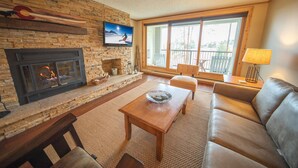 A living room with a fireplace, a television mounted on the wall, a sofa, a coffee table, and a balcony with a view. - Pines 2116 (Keystone)