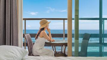 A room with a large window, a bed, a wicker chair, and a view of the ocean.