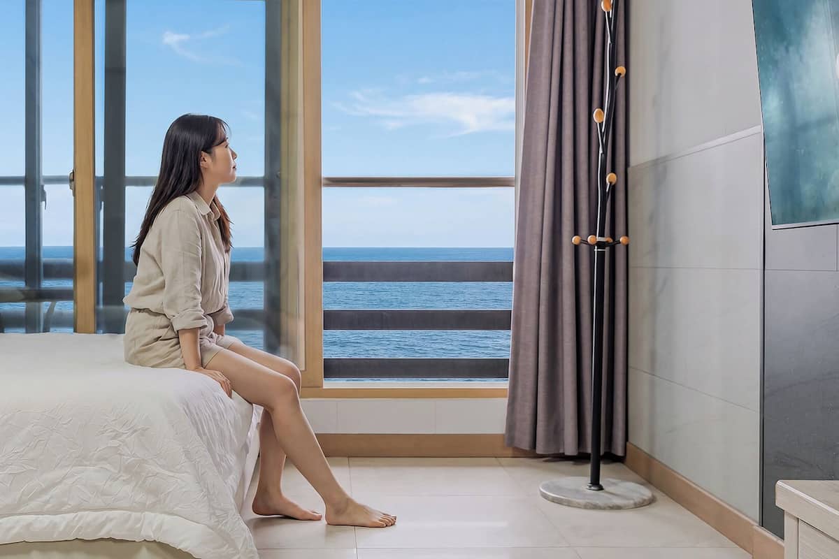 Una habitación con un amplio ventanal con vistas al mar, una cama con colcha blanca y una lámpara de pie.