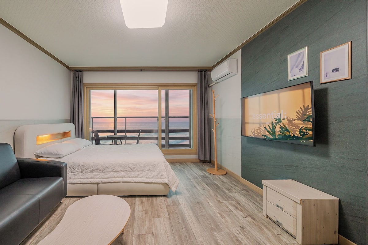 Habitación de hotel con cama, sofá, una mesita, televisor y vista al océano al atardecer.
