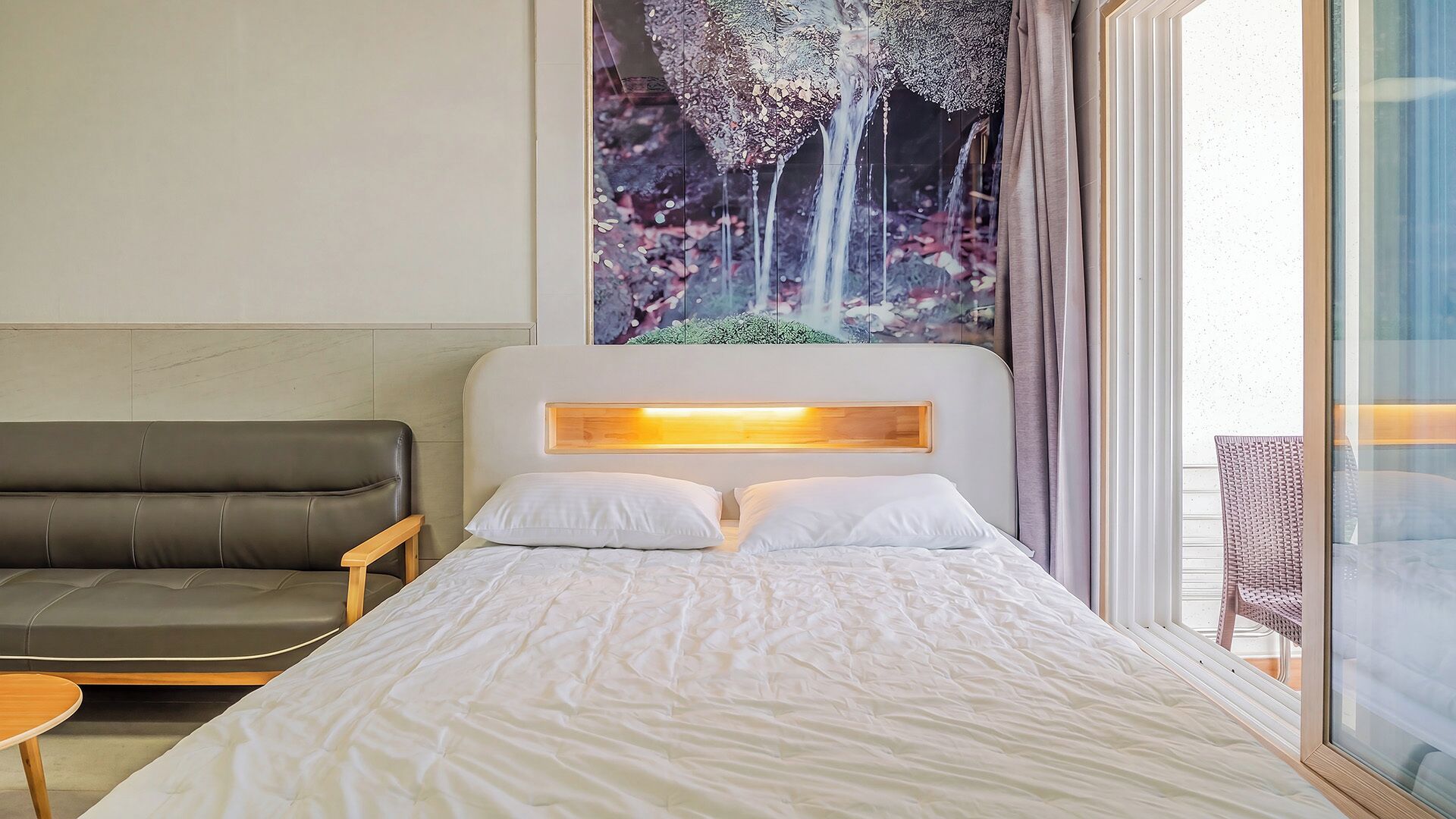 A hotel room with a bed, a sofa, a wooden side table, and a large window with a view of trees.
