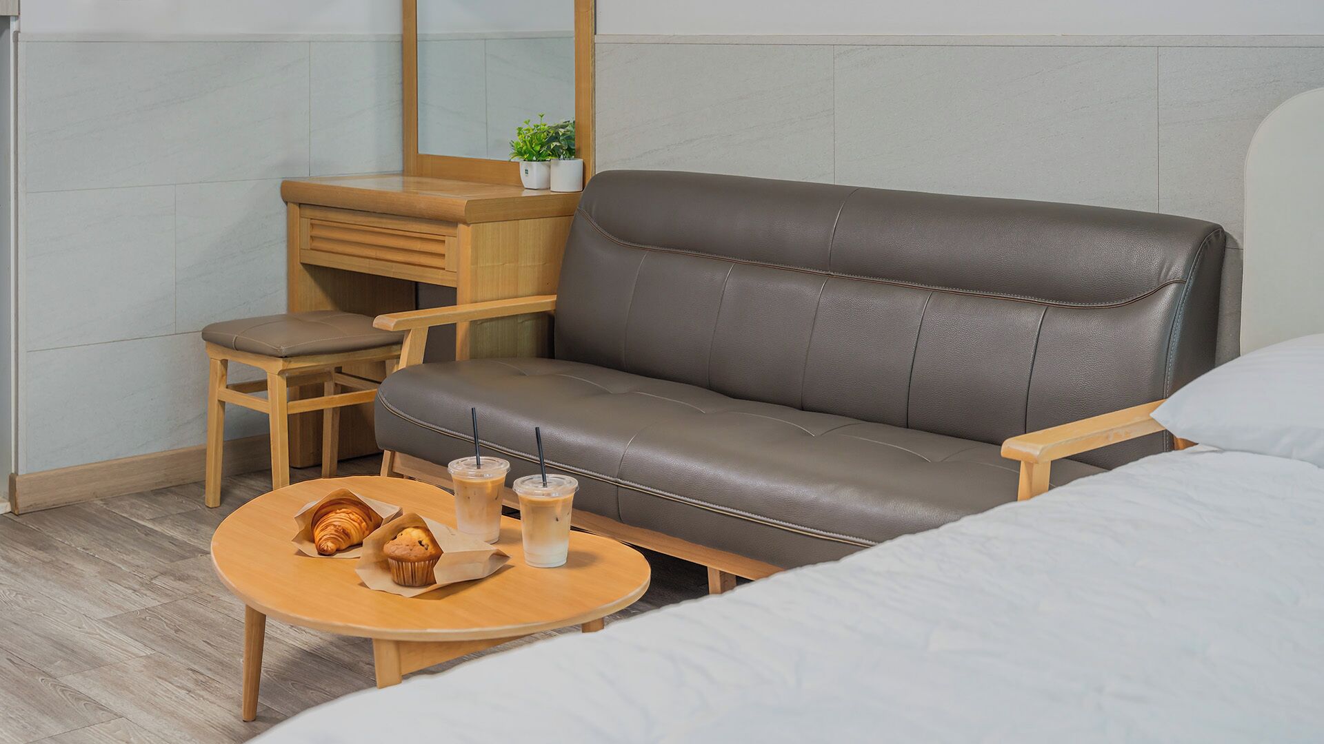 A modern hotel room with a leather sofa, a wooden desk, a small round table with pastries and drinks, and a bed with white bedding.