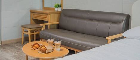 A modern hotel room with a leather sofa, a wooden desk, a small round table with pastries and drinks, and a bed with white bedding.