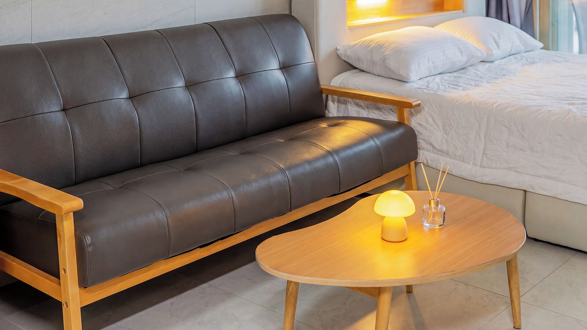 A modern room with a leather sofa, a wooden coffee table with a lamp, and a bed with white bedding.