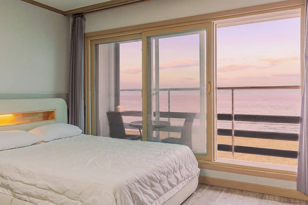 Habitación de hotel con cama, una silla, una mesa y un amplio ventanal corredero que da a un balcón con vista al mar.