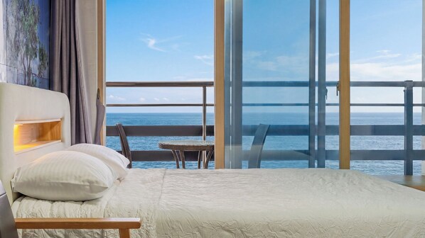 Habitación de hotel con una cama grande, una mesita pequeña y vistas al mar a través de un balcón de cristal.