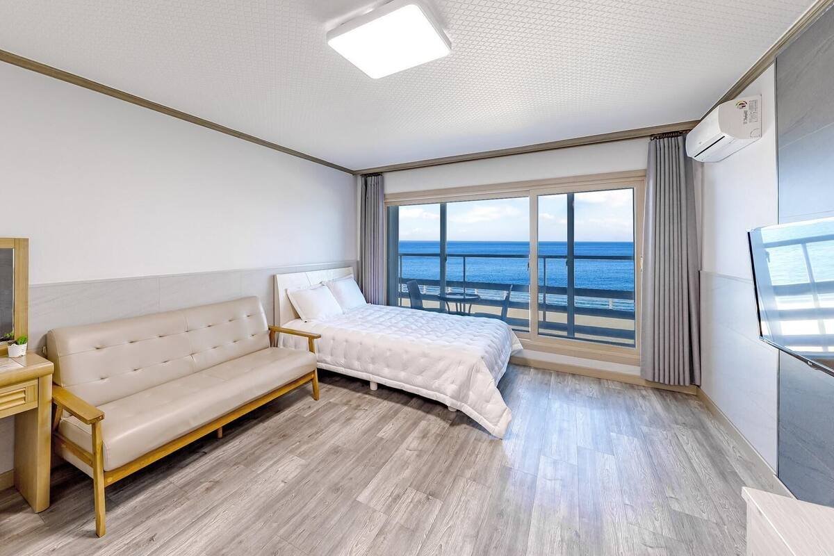 Habitación de hotel con cama, sofá, escritorio, espejo y un gran ventanal con vistas al mar.