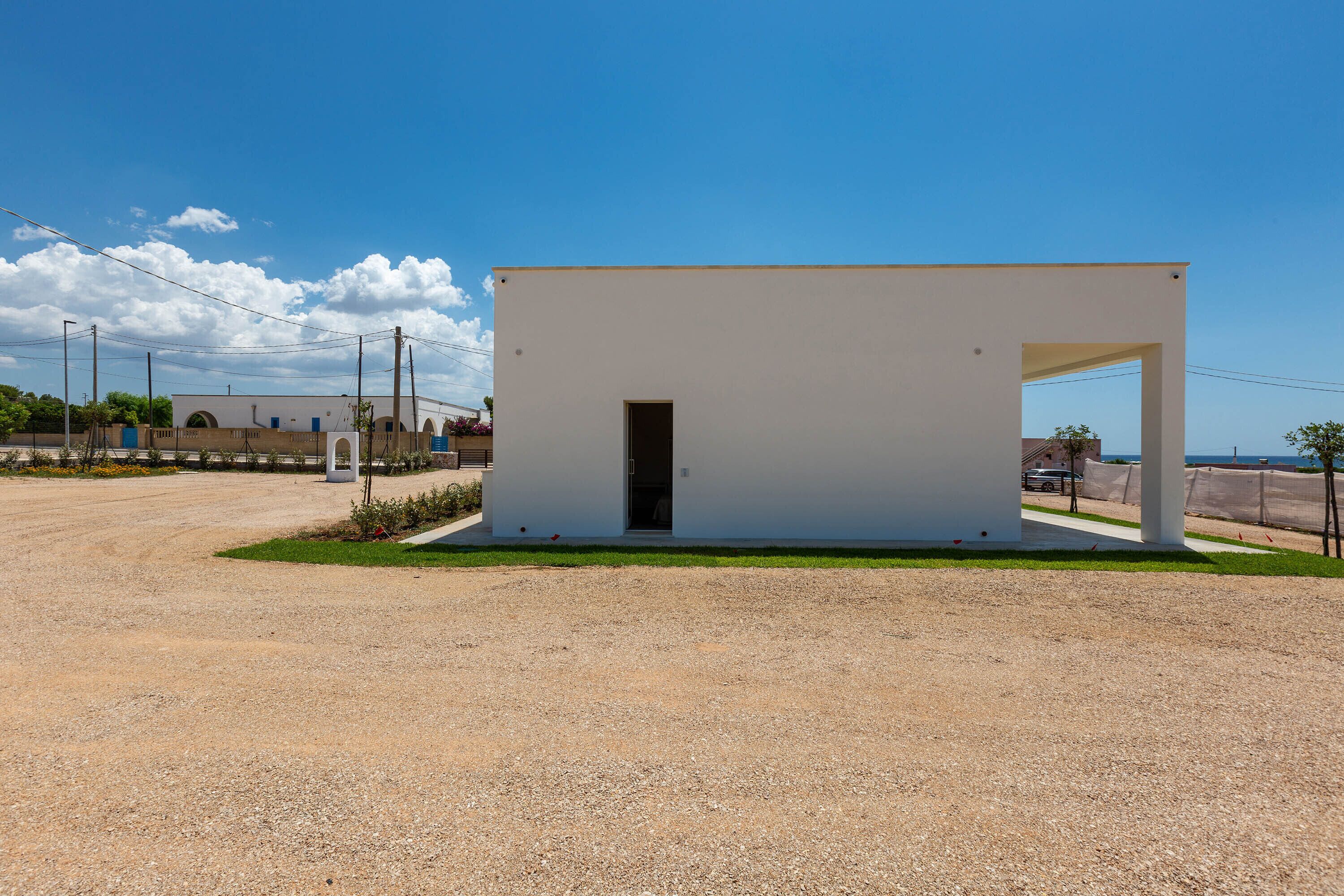 Un pequeño edificio blanco de una planta, con una puerta y un arco abierto, rodeado de grava y un cielo despejado.