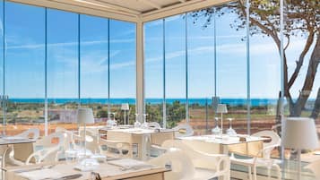 A restaurant with tables set for dining, large windows offering a view of the ocean, and a tree visible outside.