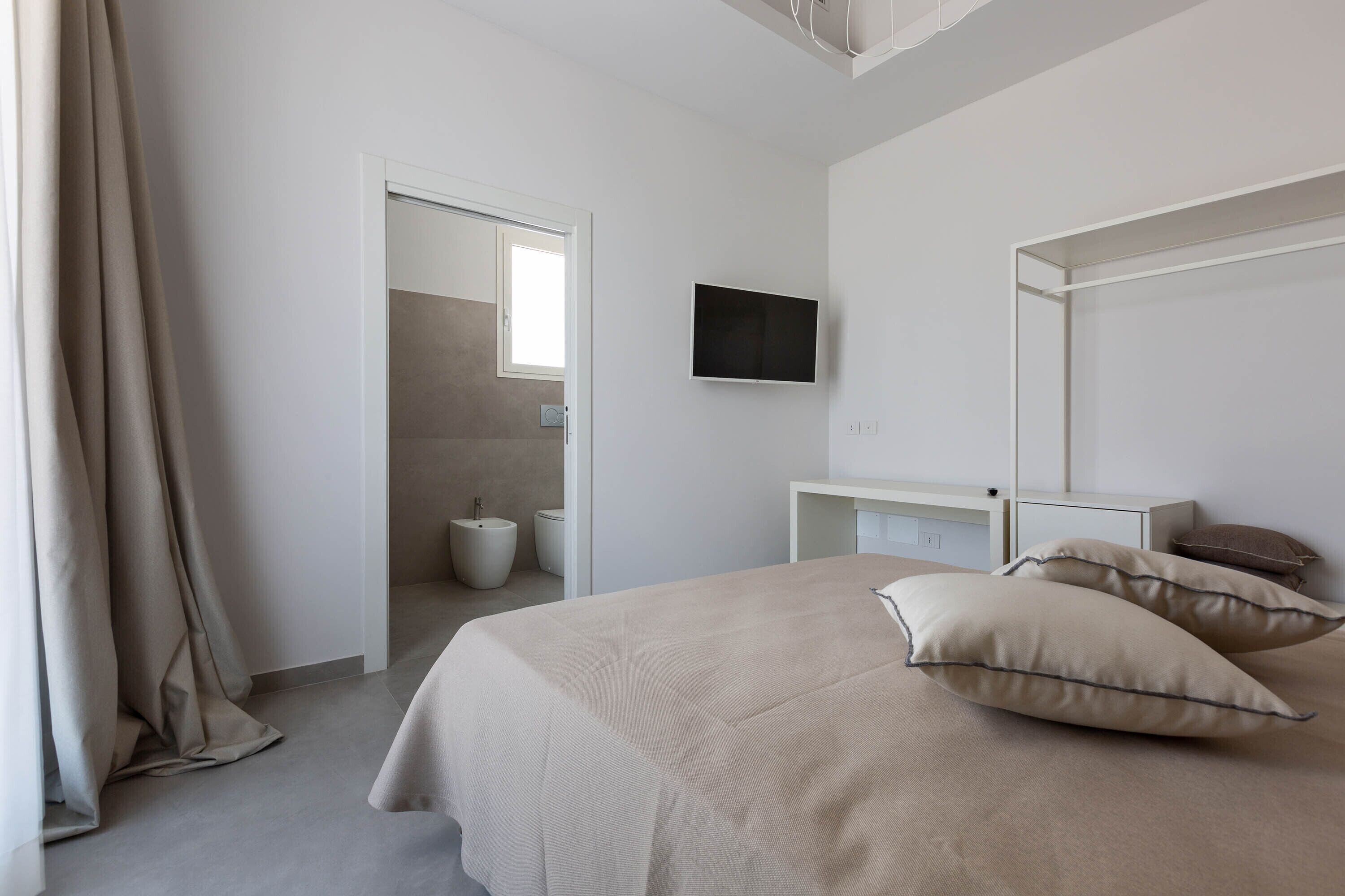 A modern bedroom with a bed, a TV, a bedside table, and a bathroom visible through an open door.