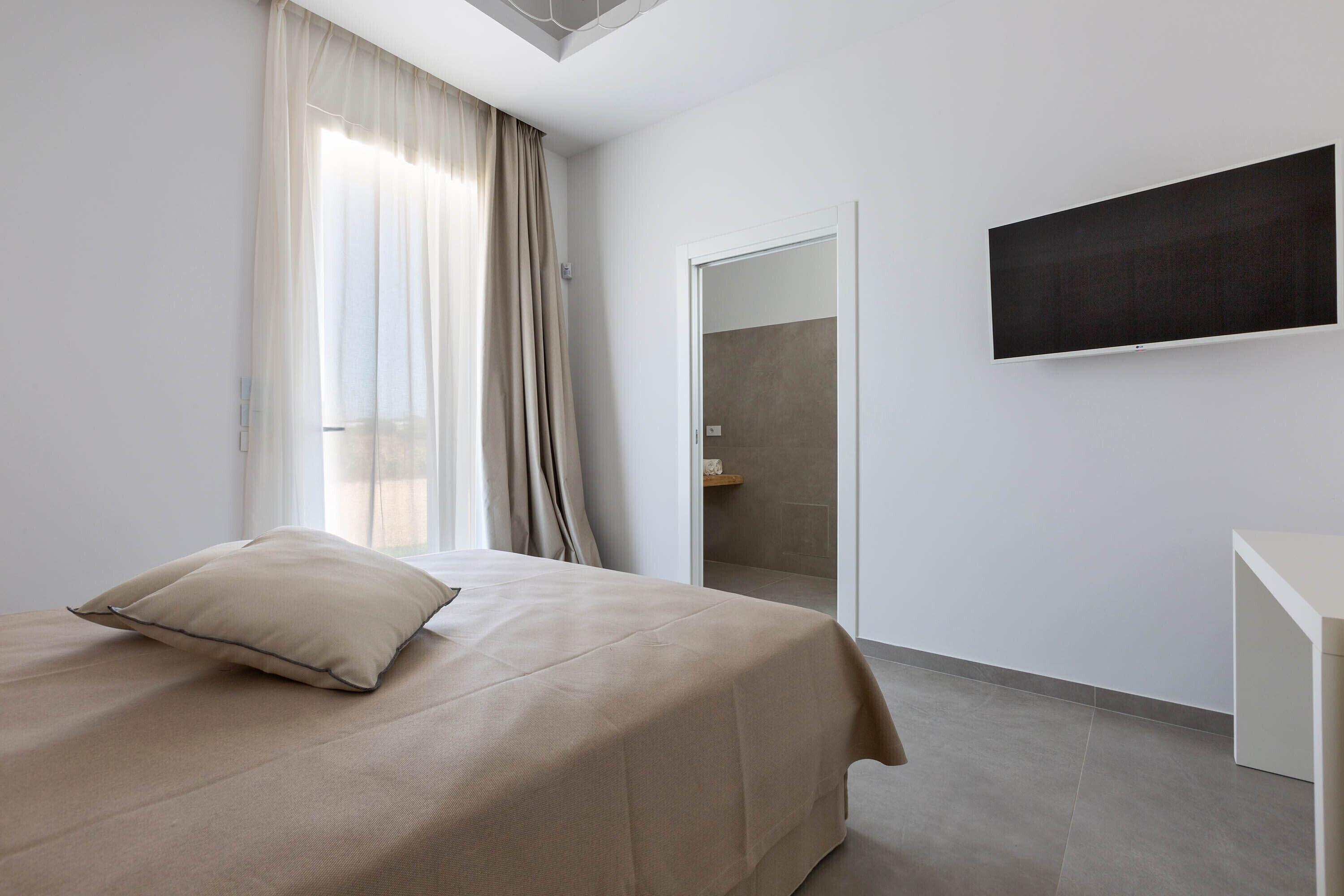 Habitación de hotel moderna con cama, televisión de pantalla plana, puerta que da a un baño y un ventanal con cortinas.