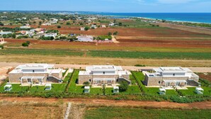 A modern residential area with three detached houses featuring stone exteriors and flat roofs, surrounded by well-maintained gardens and a coastal view.