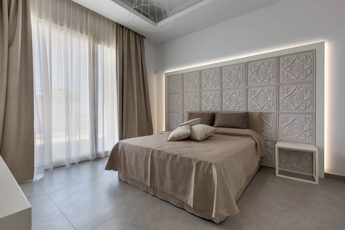 Une chambre à coucher moderne avec un grand lit, une tête de lit décorative et des rideaux légers.