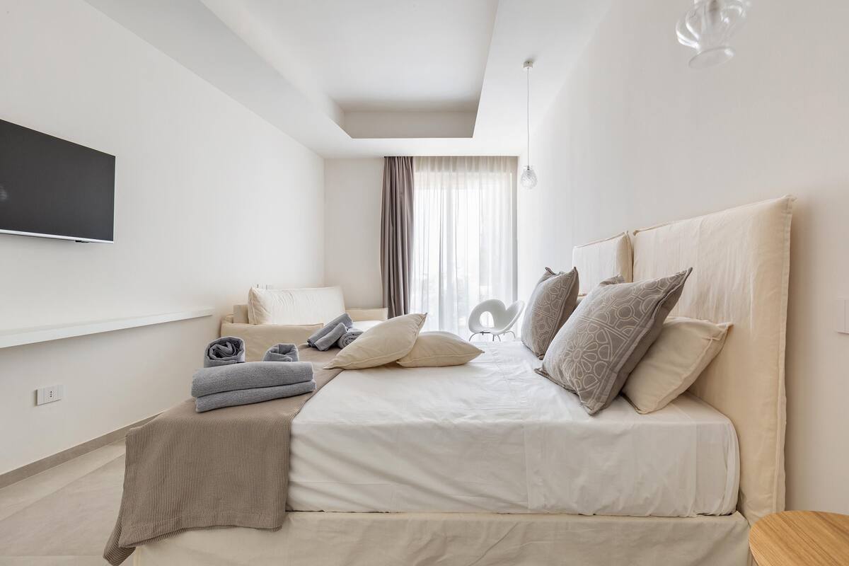 Ein modernes Hotelzimmer mit einem großen Bett, einem flachbildfernseher an der Wand, einer Sitzecke mit einer Couch und Sesseln sowie einem großen Fenster mit Vorhängen.