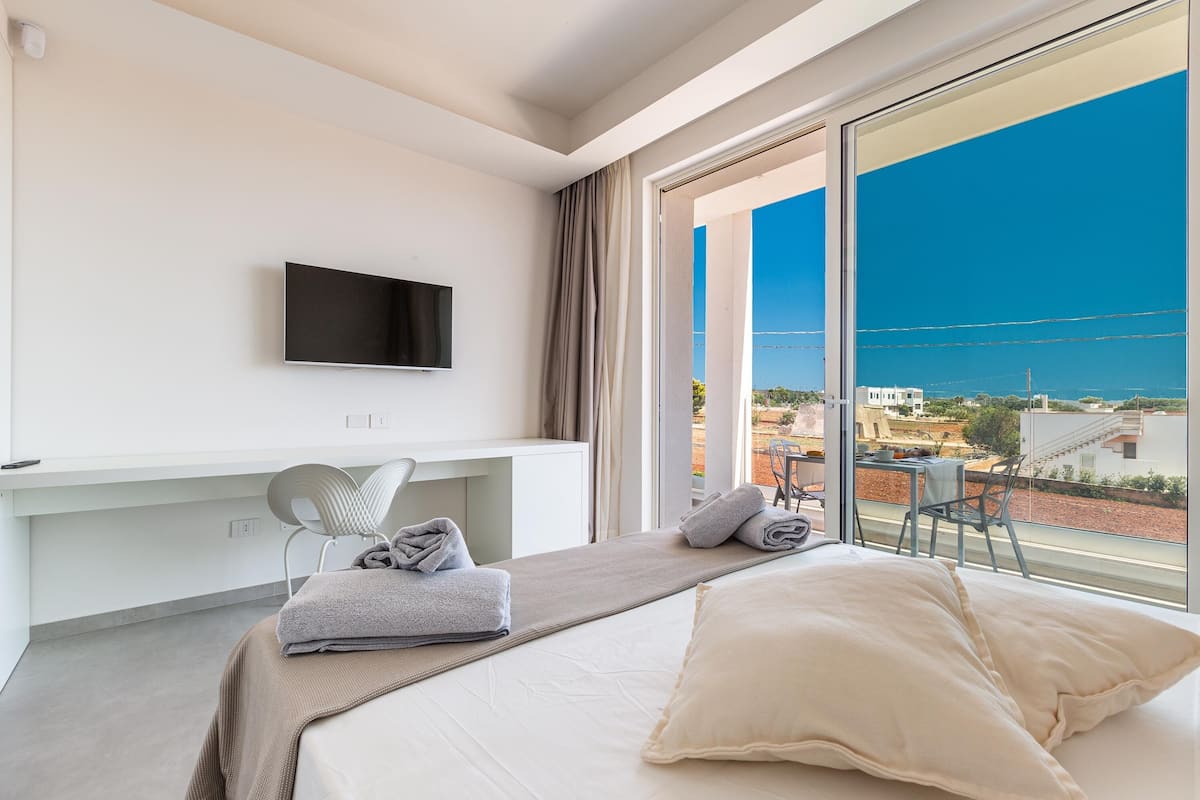 Une chambre d’hôtel avec un lit, un téléviseur fixé au mur, un bureau et une vue sur la ville grâce à de grandes fenêtres.