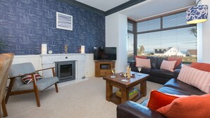 A living room with a fireplace, a television, and a sofa with red cushions. - Oystercatcher House (St Ives)