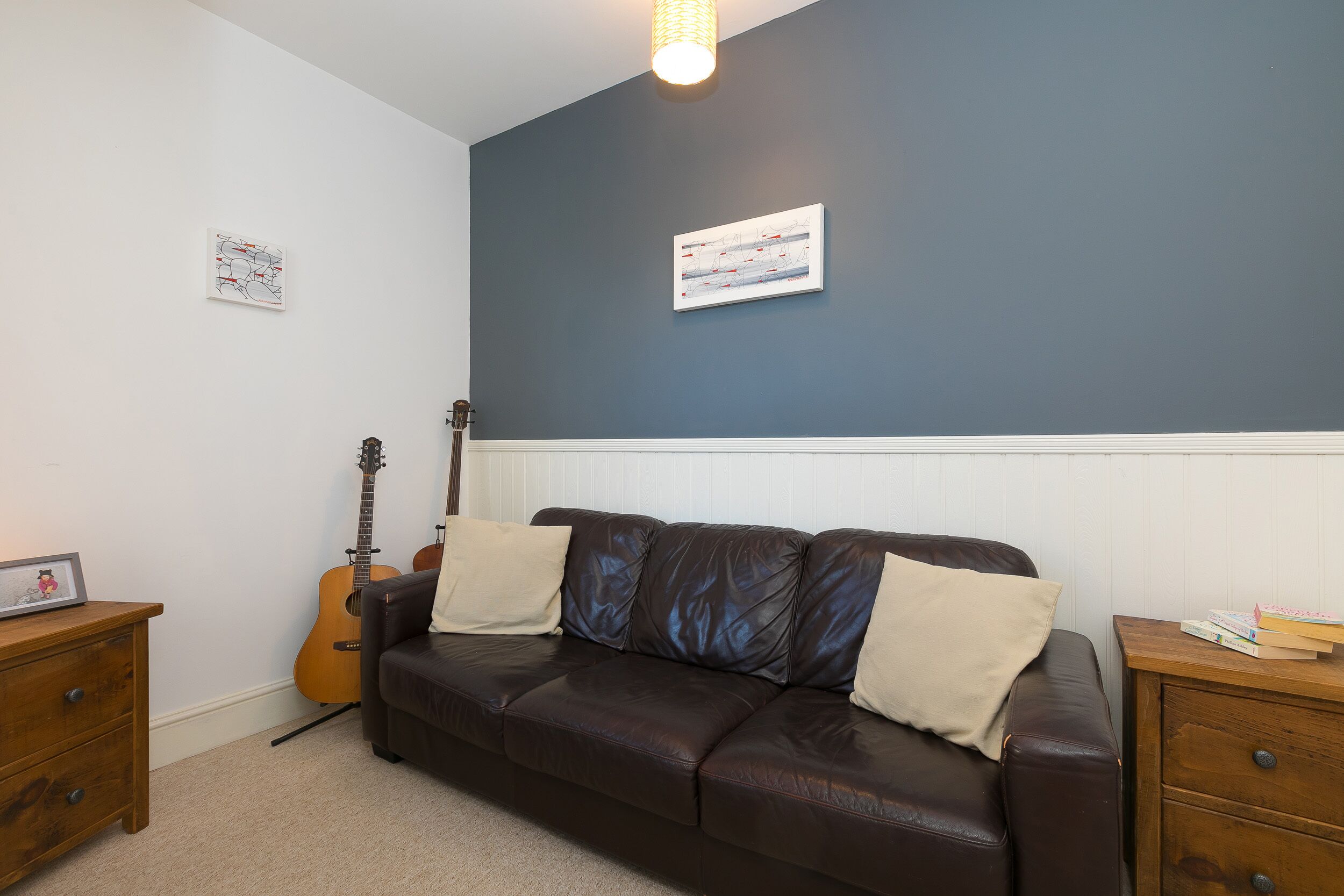 Una habitación con un sofá de cuero marrón, una guitarra apoyada en la pared y una mesita de noche de madera con libros.