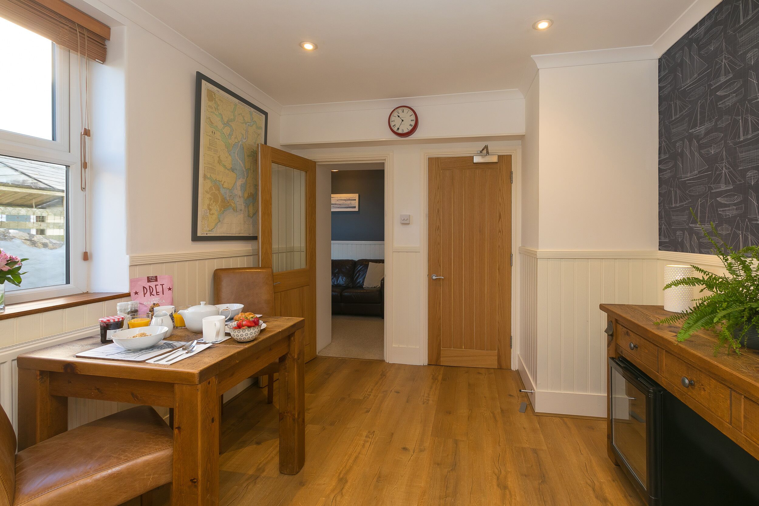 A dining area with a wooden table set for breakfast, a clock on the wall, and a view into a living room through an open door.